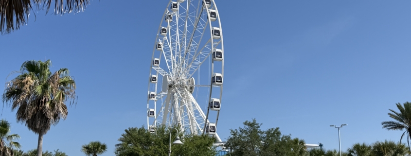 A high-angle view of the massive 200-foot PCB Wheel at Pier Park in Panama City Beach, featuring its fully enclosed, climate-controlled gondolas against a bright blue Florida sky.