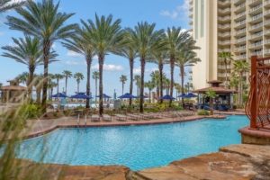 A massive tropical lagoon-style resort pool surrounded by tall palm trees and high-rise condo buildings under a clear blue sky at Shores of Panama in Panama City Beach, Florida.