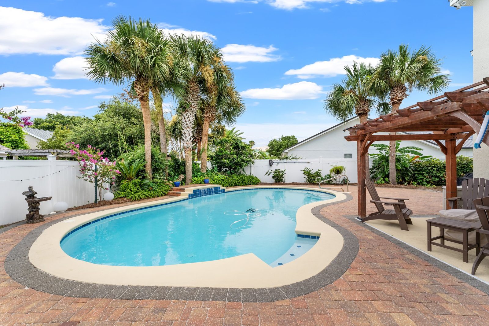 Backyard with a curved private pool, brick paver deck, palm trees, and a wooden pergola with seating